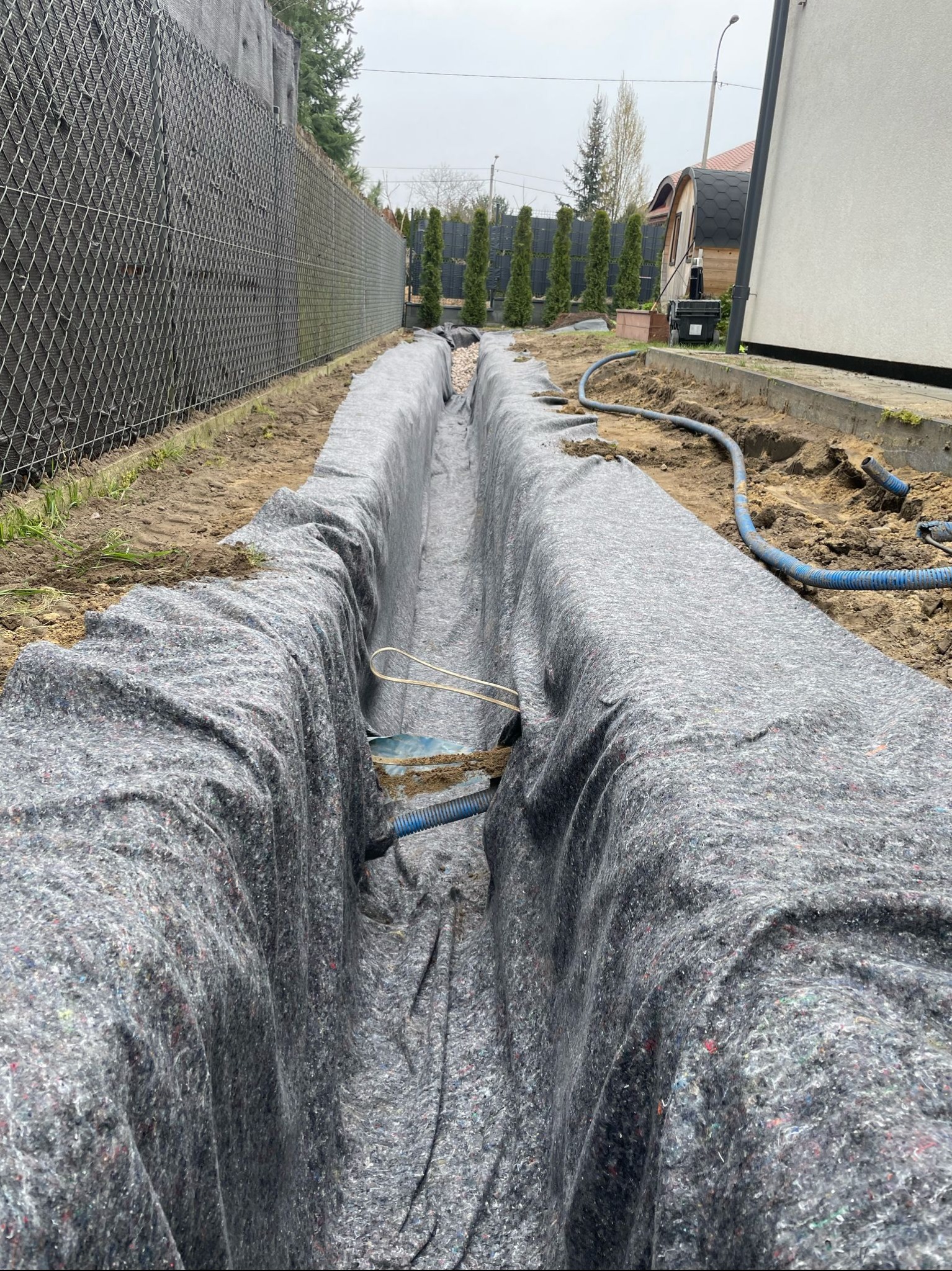Wykop liniowy zabezpieczony szarą włókniną, widoczne rury instalacyjne. W tle ogrodzenie z siatki i rząd tuj. Prace ziemne w ogrodzie.