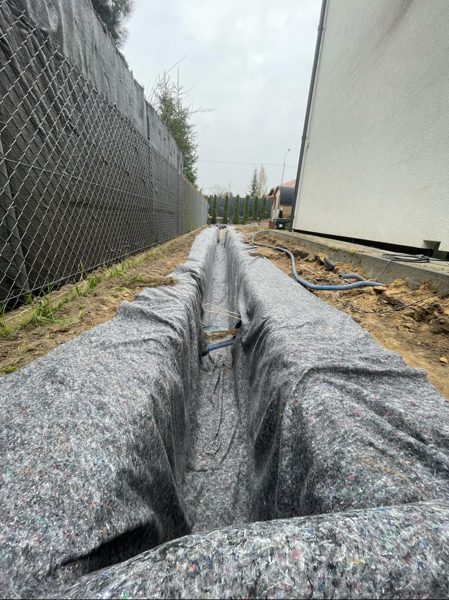 Wykop liniowy wyłożony szarą geowłókniną, prawdopodobnie pod instalacje, widoczny przewód. W tle ogrodzenie z siatki i ściana budynku. Pochmurny dzień.