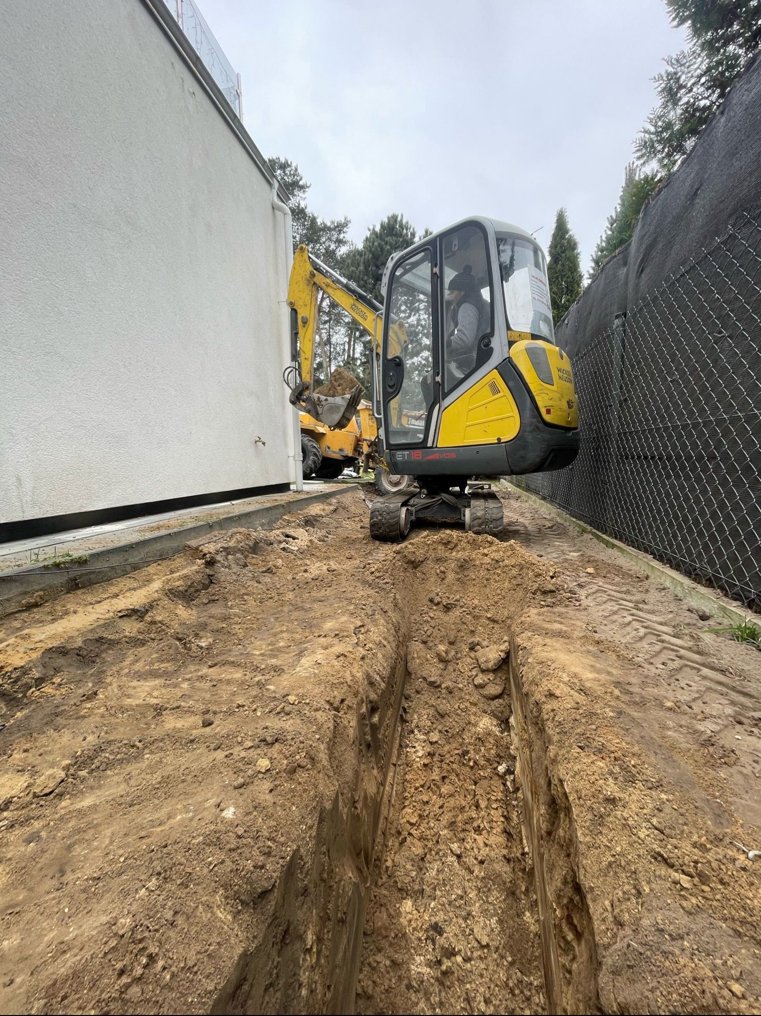 Żółta mini koparka Wacker Neuson ET18 kopie wąski wykop w ziemi wzdłuż białego budynku i ogrodzenia z siatki. Widoczny operator w kabinie.