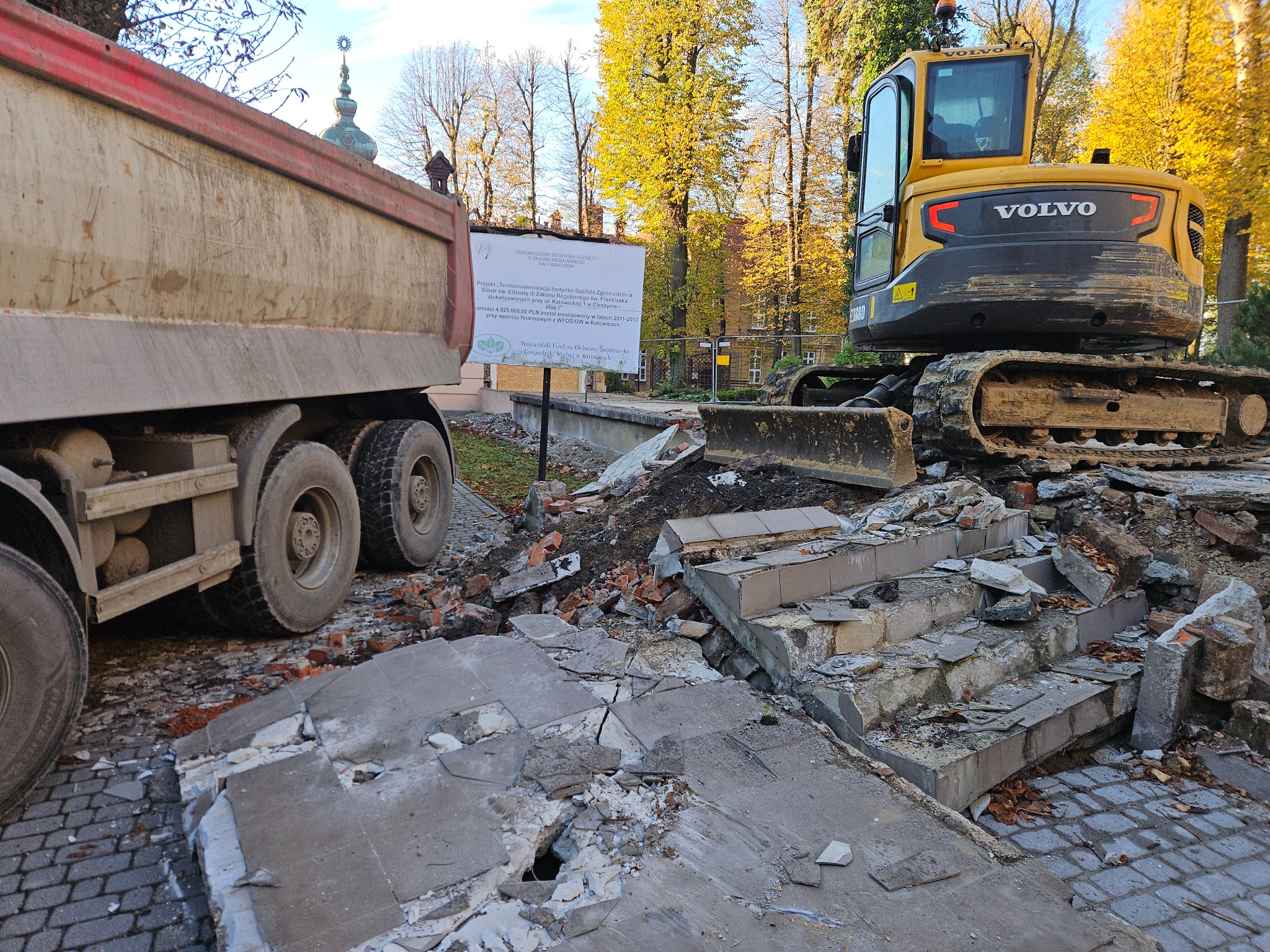 Żółta koparka Volvo na gruzowisku po rozbiórce schodów, obok brudna ciężarówka, w tle budynek z wieżyczką i drzewa w jesiennych barwach.