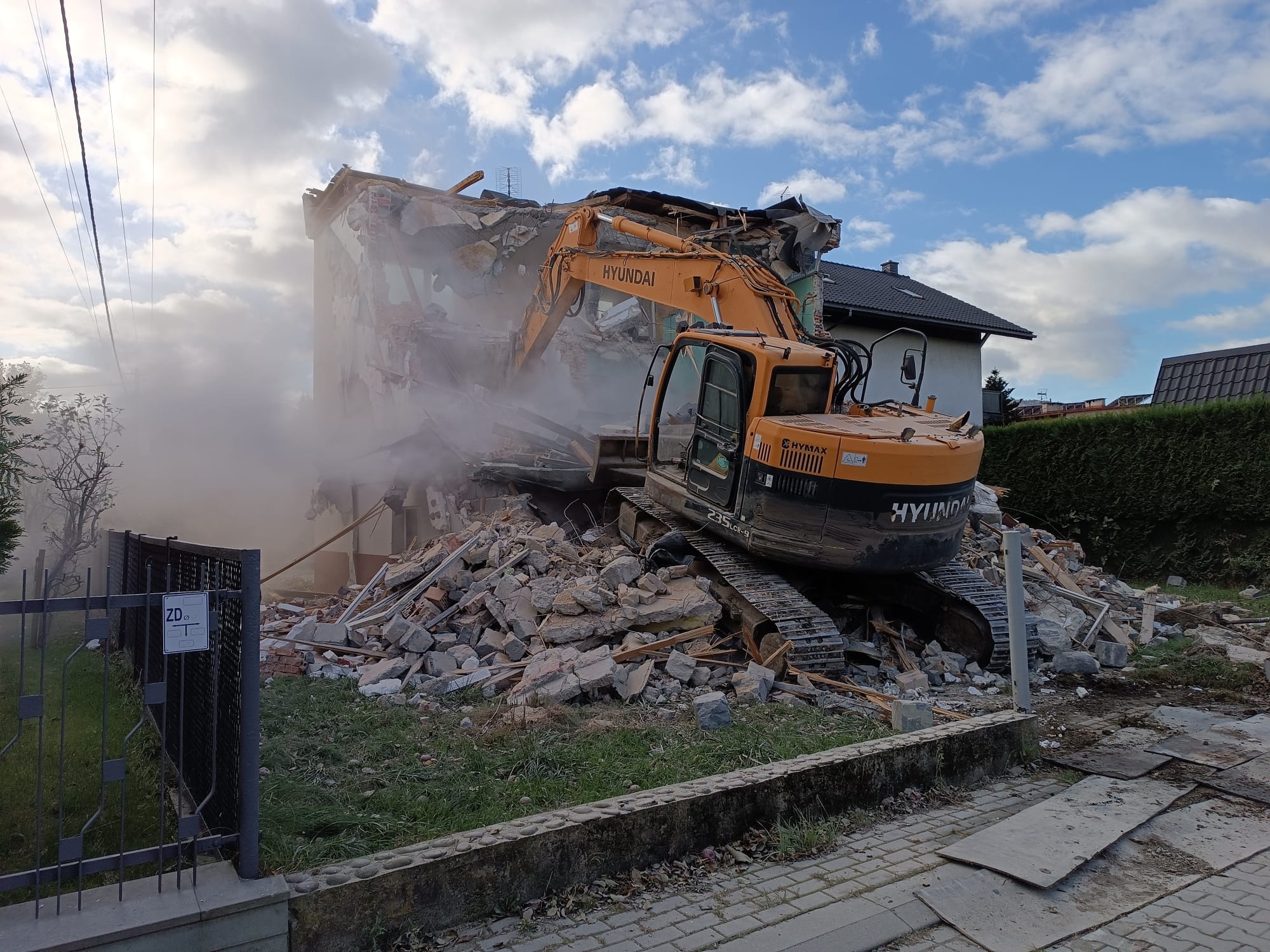 Pomarańczowa koparka Hyundai na tle częściowo zburzonego, dwupiętrowego budynku mieszkalnego; gruzowisko i tumany kurzu w powietrzu, widoczne są fragmenty dachu i okien.