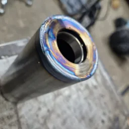 Spojrzenie z bliska na metalowy cylinder z widocznym spoiną o opalizujących barwach, leżący na warsztatowym stole.