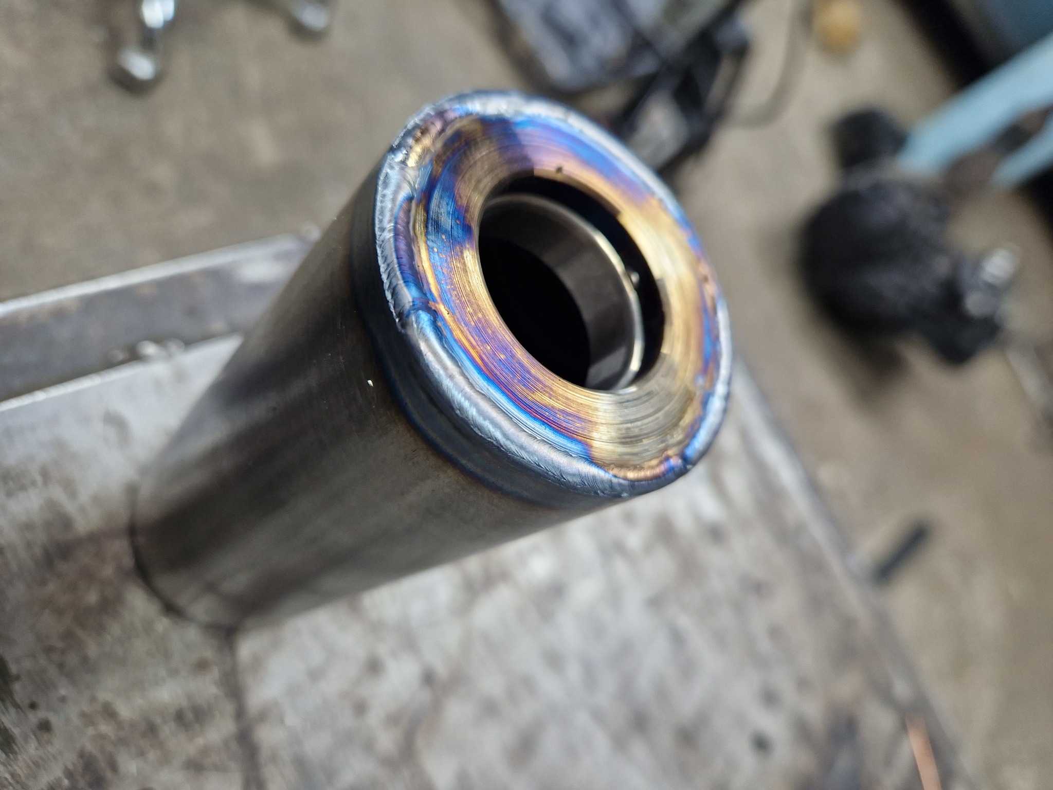 Spojrzenie z bliska na metalowy cylinder z widocznym spoiną o opalizujących barwach, leżący na warsztatowym stole.