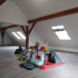 Michał Szatkowski - Poddasze w trakcie prac wykończeniowych: widoczne drewniane belki konstrukcyjne, okna dachowe, podłoga z paneli, stół z narzędziami, materiałami budowlanymi i napojami, rozłożona czerwona folia...