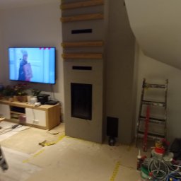 Michał Szatkowski - Wnętrze w trakcie prac: obudowa kominka z drewnianymi elementami konstrukcyjnymi, zabezpieczona podłoga, drabina oparta o ścianę na poddaszu, telewizor z włączonym filmem w tle.