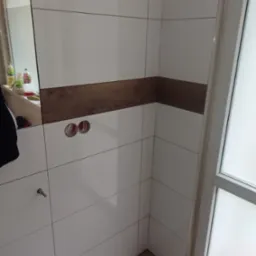 Ściana łazienki wyłożona białymi płytkami z poziomym pasem imitującym drewno, widoczne otwory na gniazdka elektryczne i fragment podłogi z paneli.