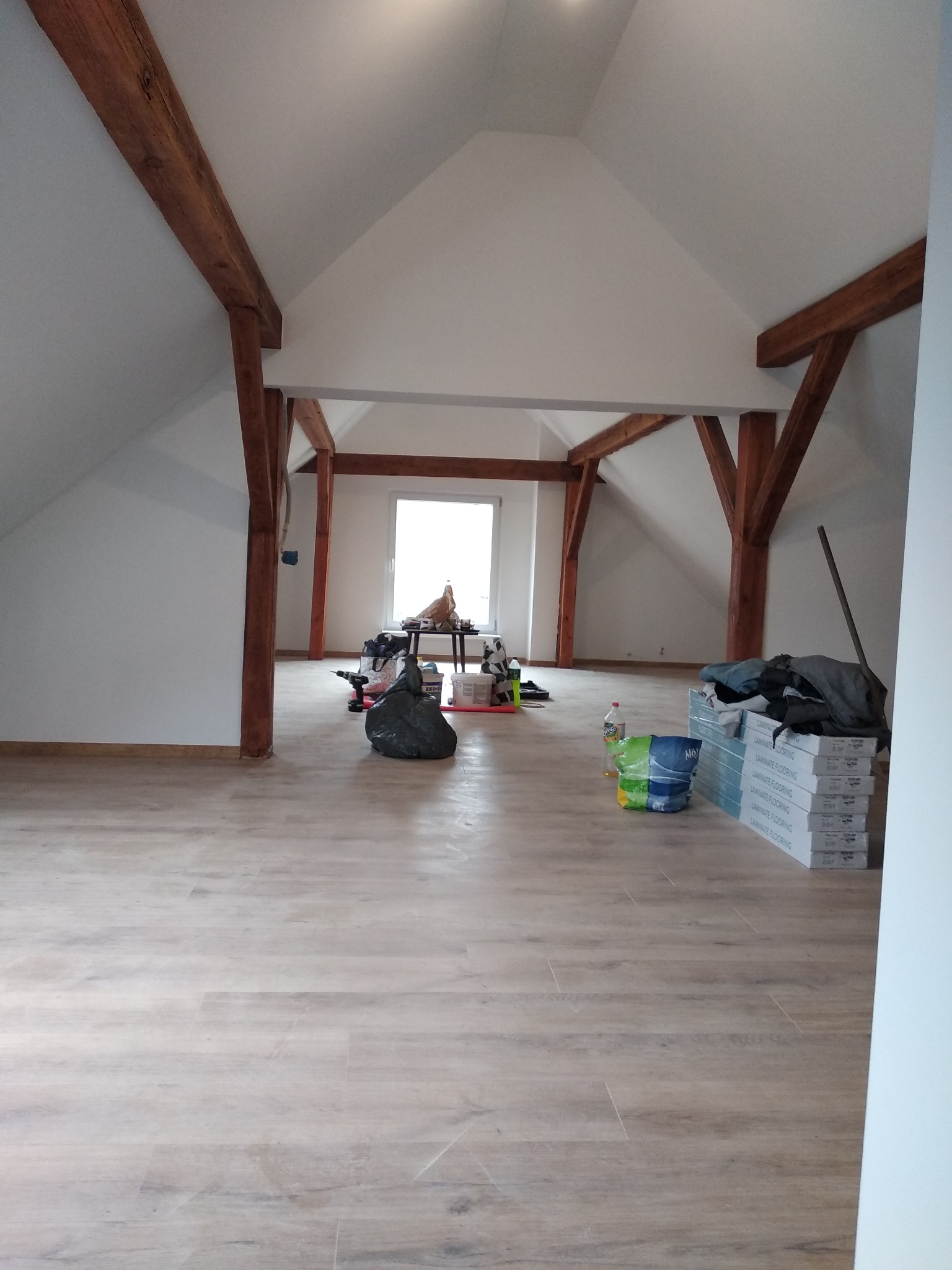 Poddasze z drewnianą konstrukcją stropu, świeżo ułożone panele podłogowe imitujące drewno, narzędzia i materiały budowlane w trakcie prac wykończeniowych.