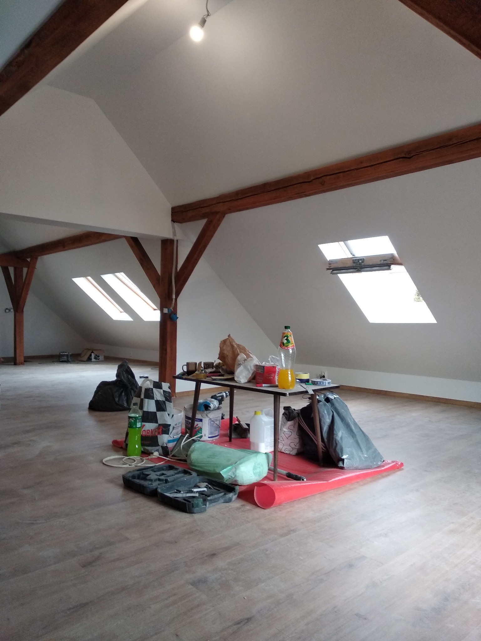 Poddasze w trakcie prac wykończeniowych: widoczne drewniane belki konstrukcyjne, okna dachowe, podłoga z paneli, stół z narzędziami, materiałami budowlanymi i napojami, rozłożona czerwona folia...