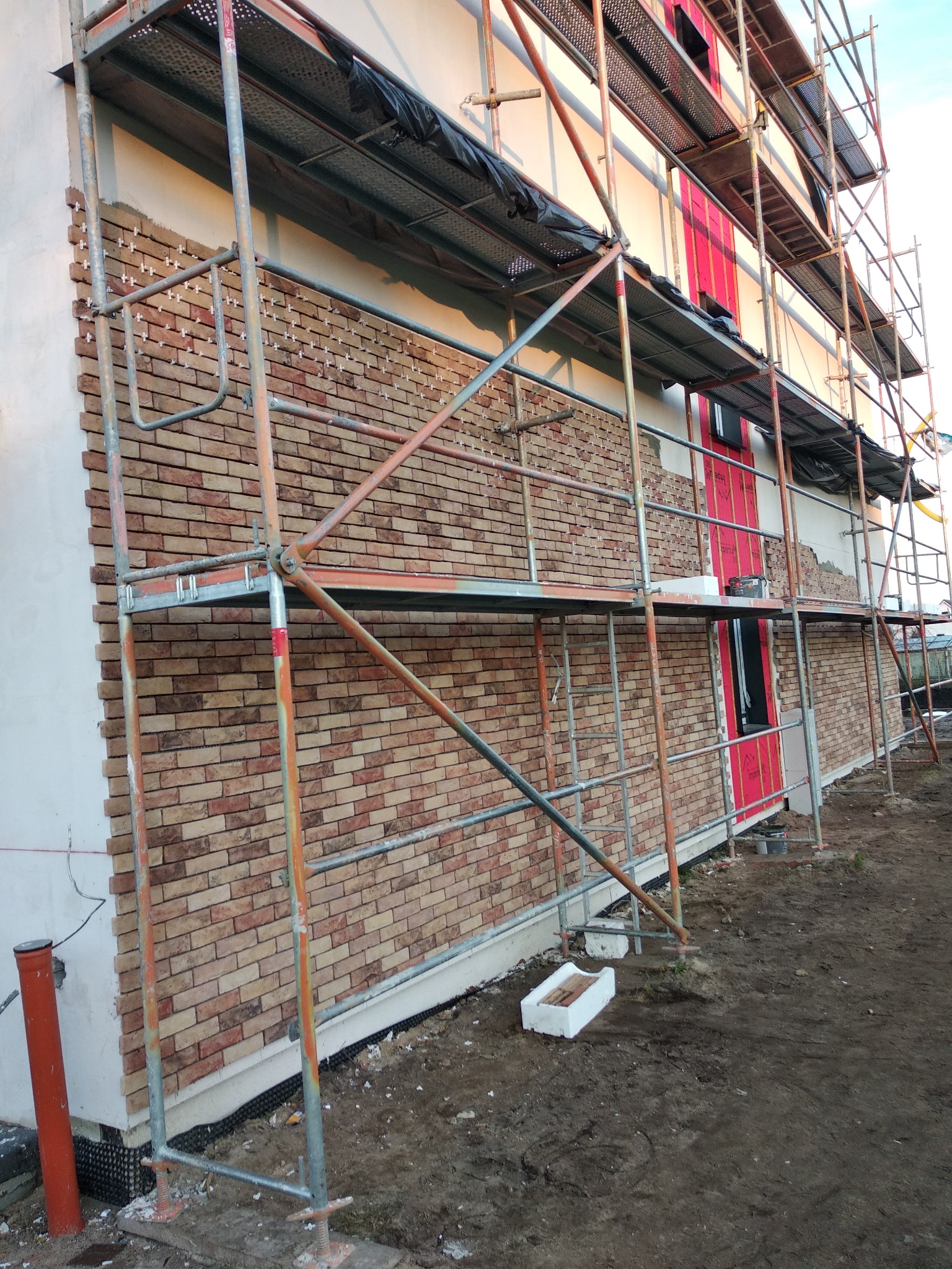 Montaż elewacji z cegły klinkierowej na budynku jednorodzinnym, widoczne rusztowania i białe krzyżyki dystansowe między cegłami.