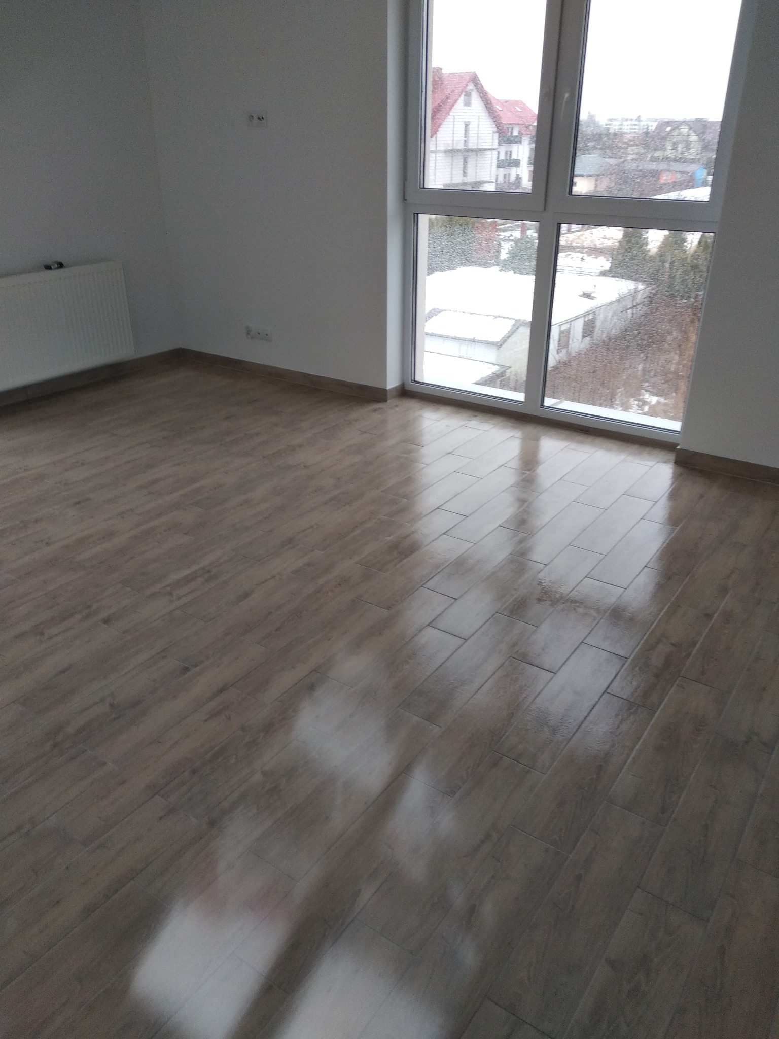 Ułożone panele podłogowe imitujące drewno w pustym pokoju z dużym oknem, za którym widać zimowy krajobraz.