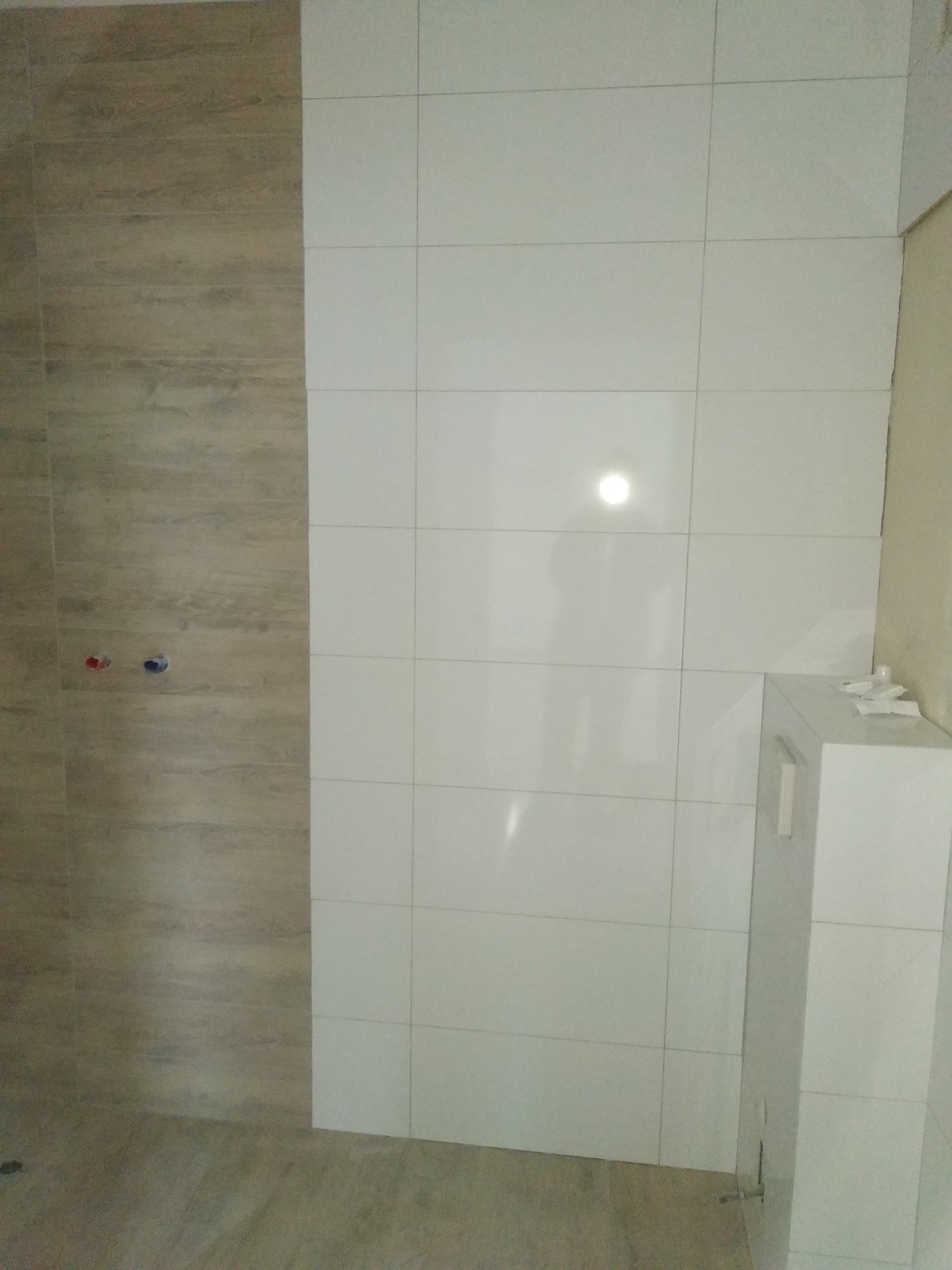 Wykończona ściana łazienki z połączeniem płytek imitujących drewno i białych kwadratowych płytek ceramicznych, widoczne przyłącza wodne i zabudowa podtynkowego WC.