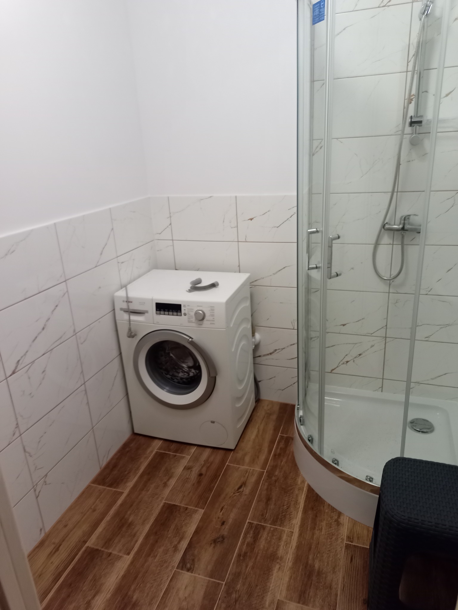 Nowoczesna łazienka z drewnopodobną podłogą, kabiną prysznicową z marmurkowymi płytkami i białą pralką z akcesoriami na górze.