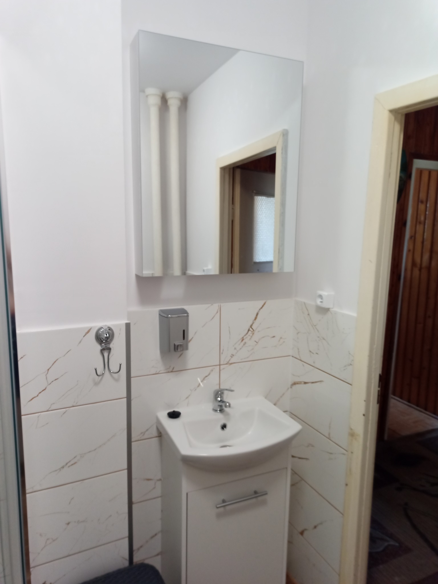 Biała umywalka z szafką i chromowaną baterią w wykończonej łazience z płytkami imitującymi marmur, widoczne akcesoria łazienkowe i lustro.