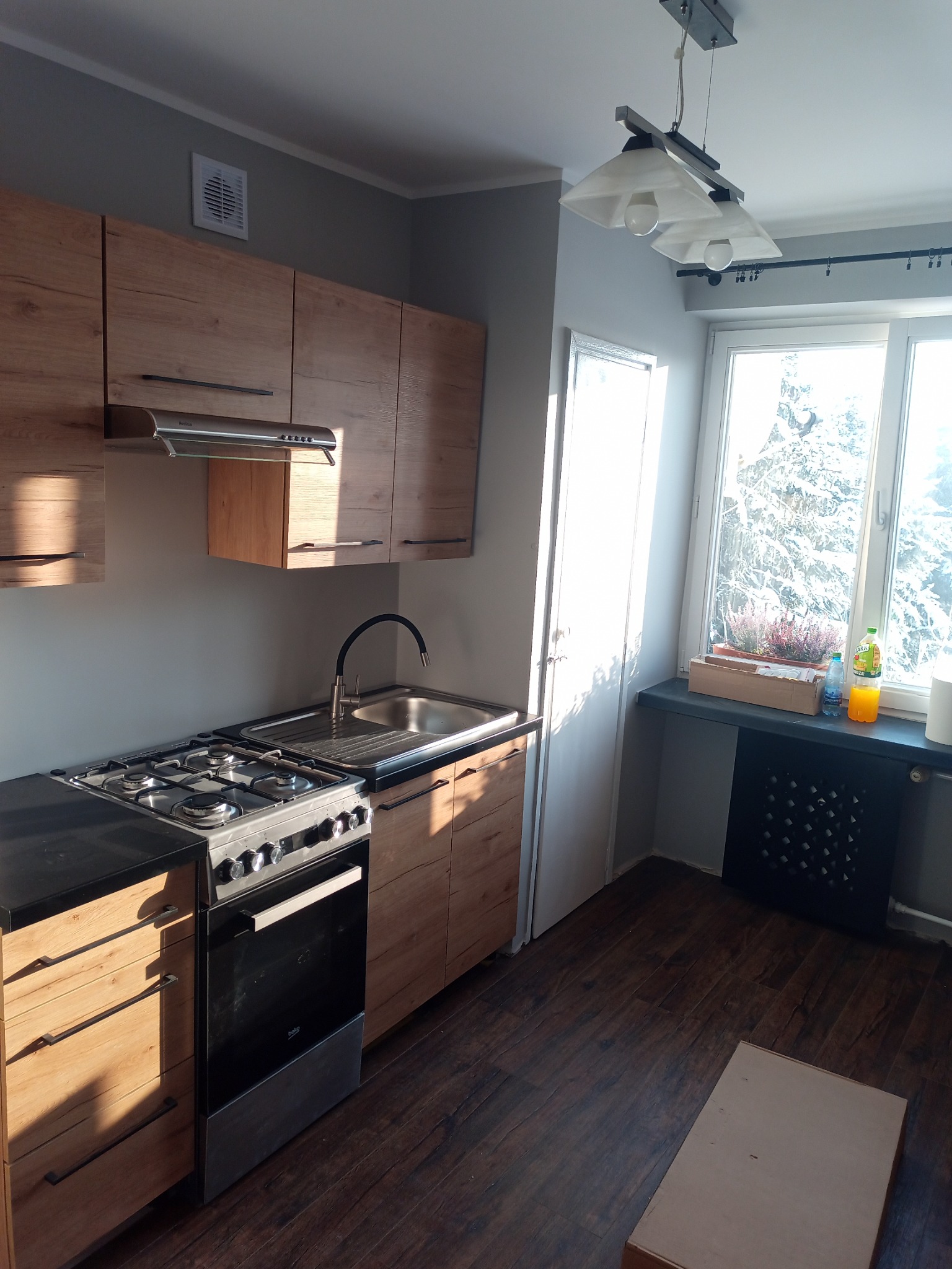 Jasna kuchnia z drewnianymi szafkami, czarnym blatem i srebrną kuchenką. Widoczne okno z roślinami i butelkami na parapecie oraz podłoga z paneli. Na podłodze kartonowe pudełko.