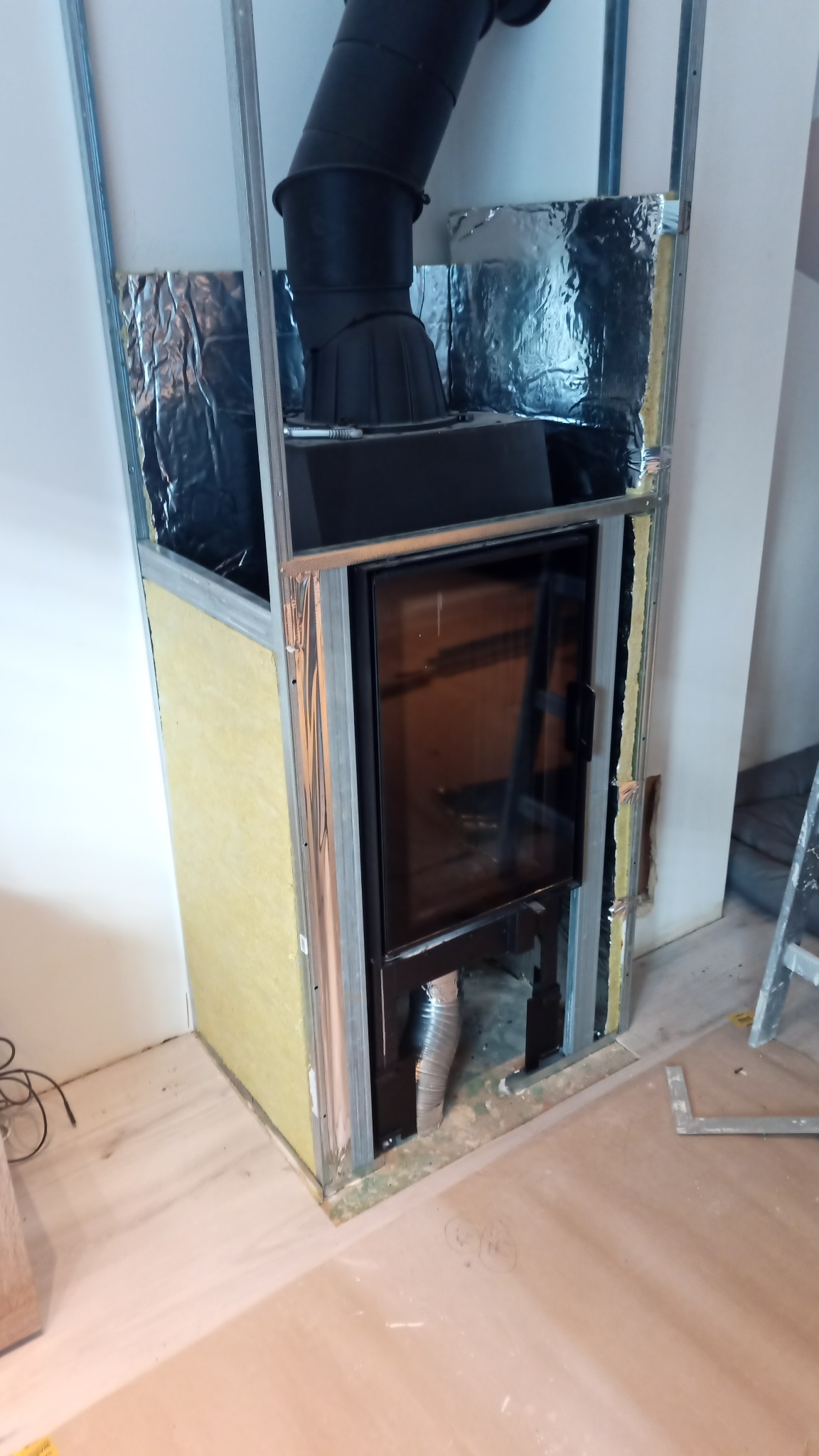 Konstrukcja obudowy kominka z płyt kartonowo-gipsowych, z izolacją z wełny mineralnej i folii aluminiowej, widoczny czarny wkład kominkowy z szybą i rurą odprowadzającą dym.