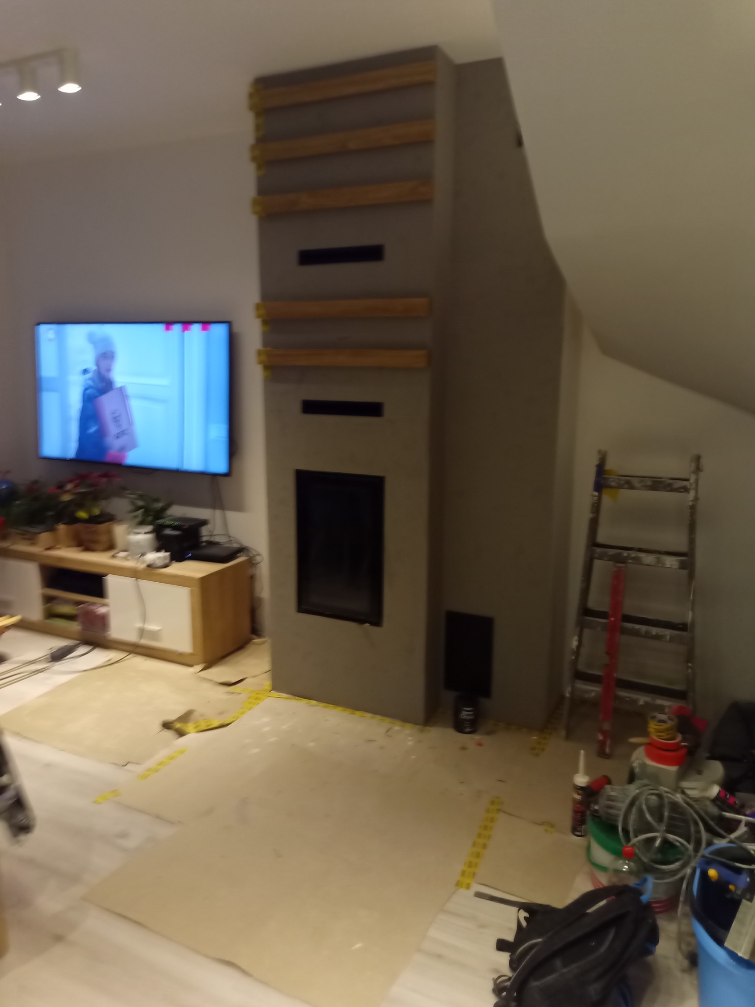 Wnętrze w trakcie prac: obudowa kominka z drewnianymi elementami konstrukcyjnymi, zabezpieczona podłoga, drabina oparta o ścianę na poddaszu, telewizor z włączonym filmem w tle.