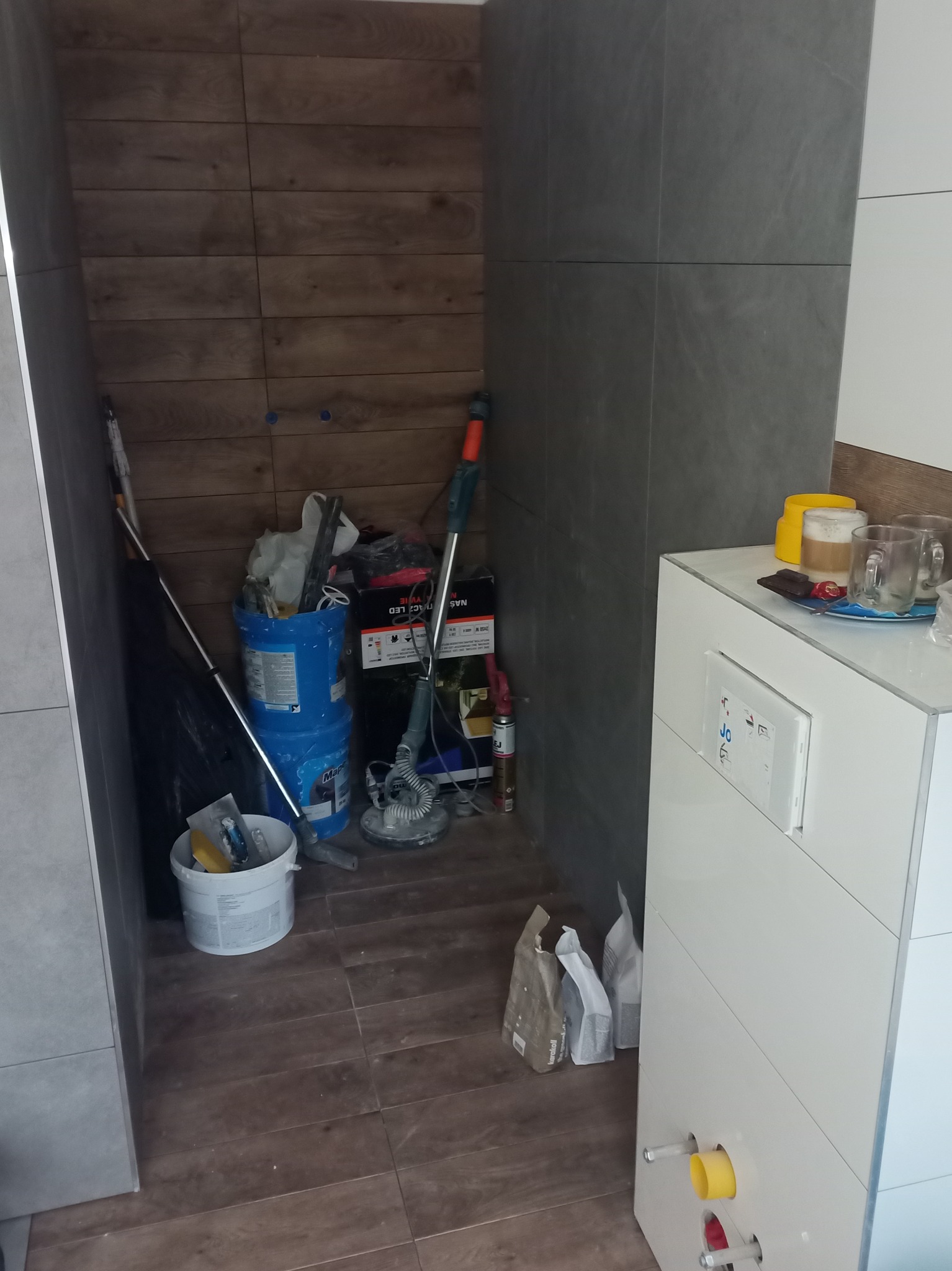 Fragment łazienki w trakcie prac wykończeniowych w Kołobrzegu, widoczne płytki imitujące drewno na ścianie, obudowa stelaża WC z przygotowanymi przyłączami, oraz narzędzia i materiały budowlane w tle.