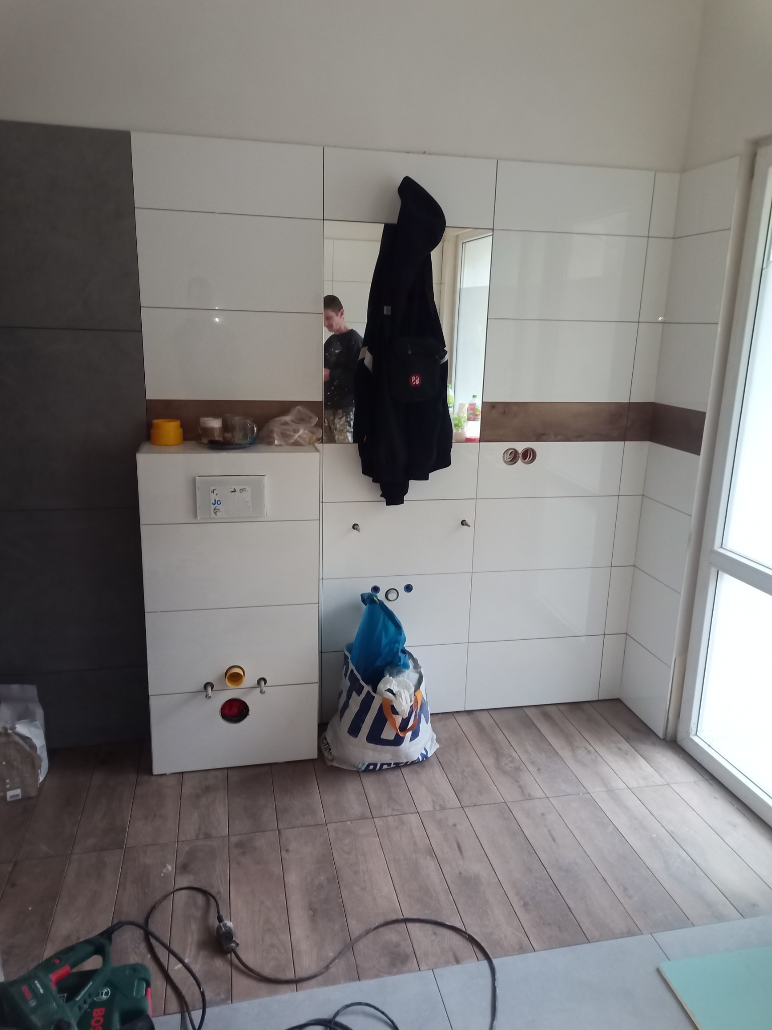 Łazienka w trakcie prac wykończeniowych: białe płytki ścienne z brązowym akcentem, podłoga wyłożona panelami imitującymi drewno, wisząca szafka z zamontowanym stelażem podtynkowym, torba...