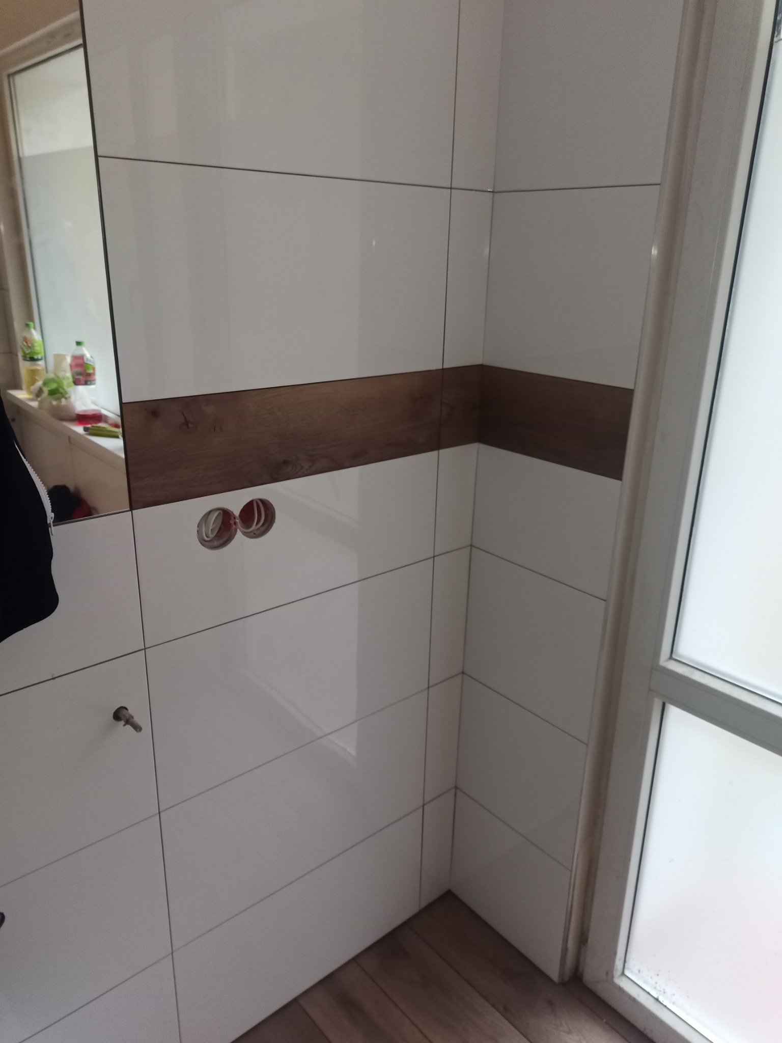 Ściana łazienki wyłożona białymi płytkami z poziomym pasem imitującym drewno, widoczne otwory na gniazdka elektryczne i fragment podłogi z paneli.