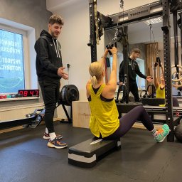 Treningi personalne Karol Kusiak - Kobieta ćwiczy na siłowni pod okiem mężczyzny, używając linki przymocowanej do sufitu, widoczne sportowe buty Adidas z turkusowymi sznurówkami.