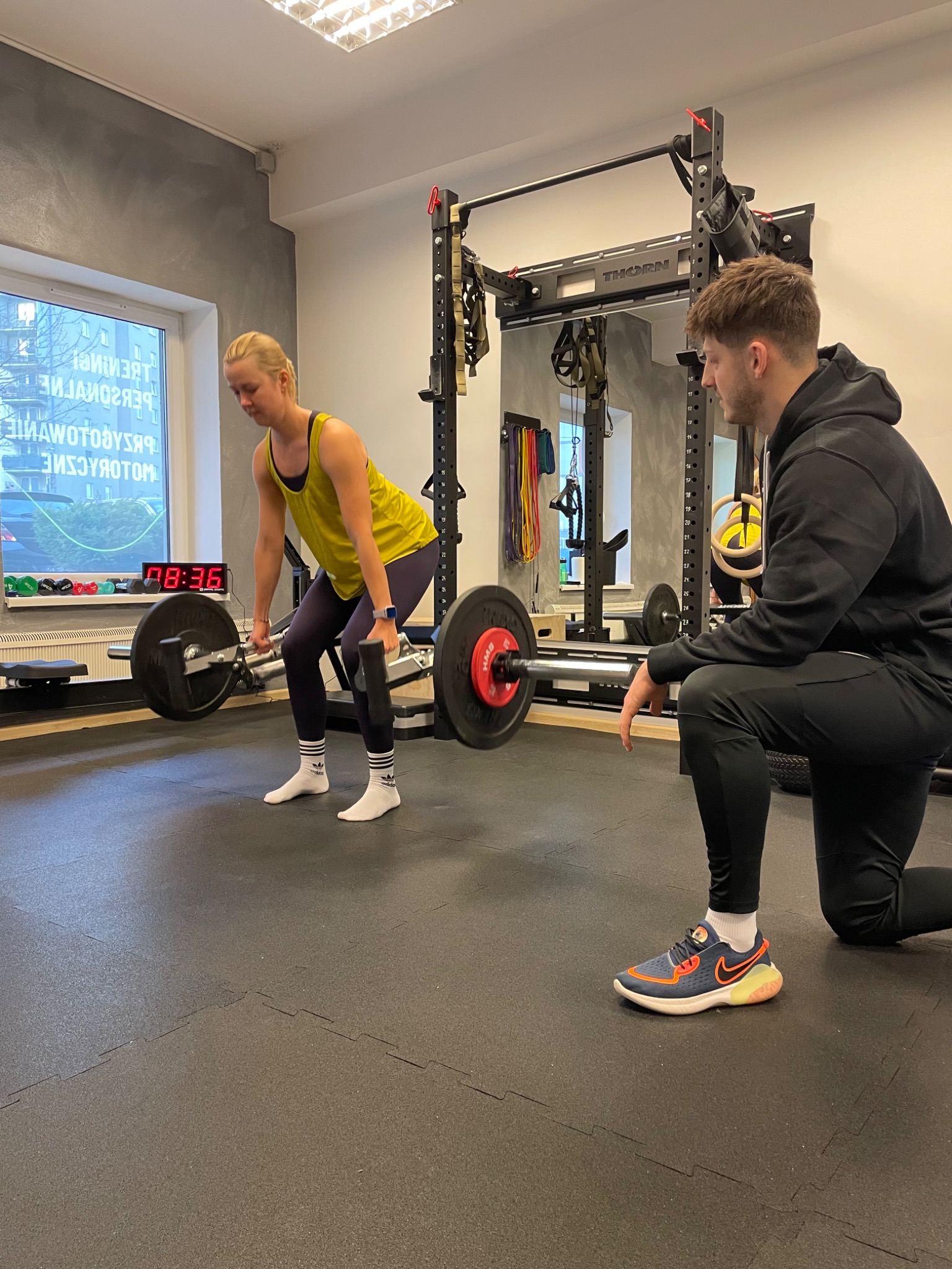 Kobieta w żółtym topie podnosi sztangę pod okiem trenera w czarnej bluzie, widoczny zegar z czasem 08:36, siłownia z profesjonalnym sprzętem.
