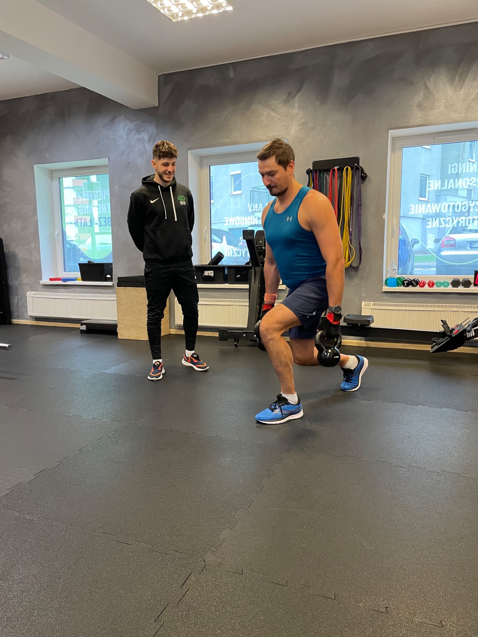 Mężczyzna w niebieskim tank topie i szortach wykonuje wypad z dwoma kettlebellami, nadzorowany przez uśmiechniętego mężczyznę w czarnej bluzie z kapturem w siłowni z gumową podłogą i szarą ścianą.
