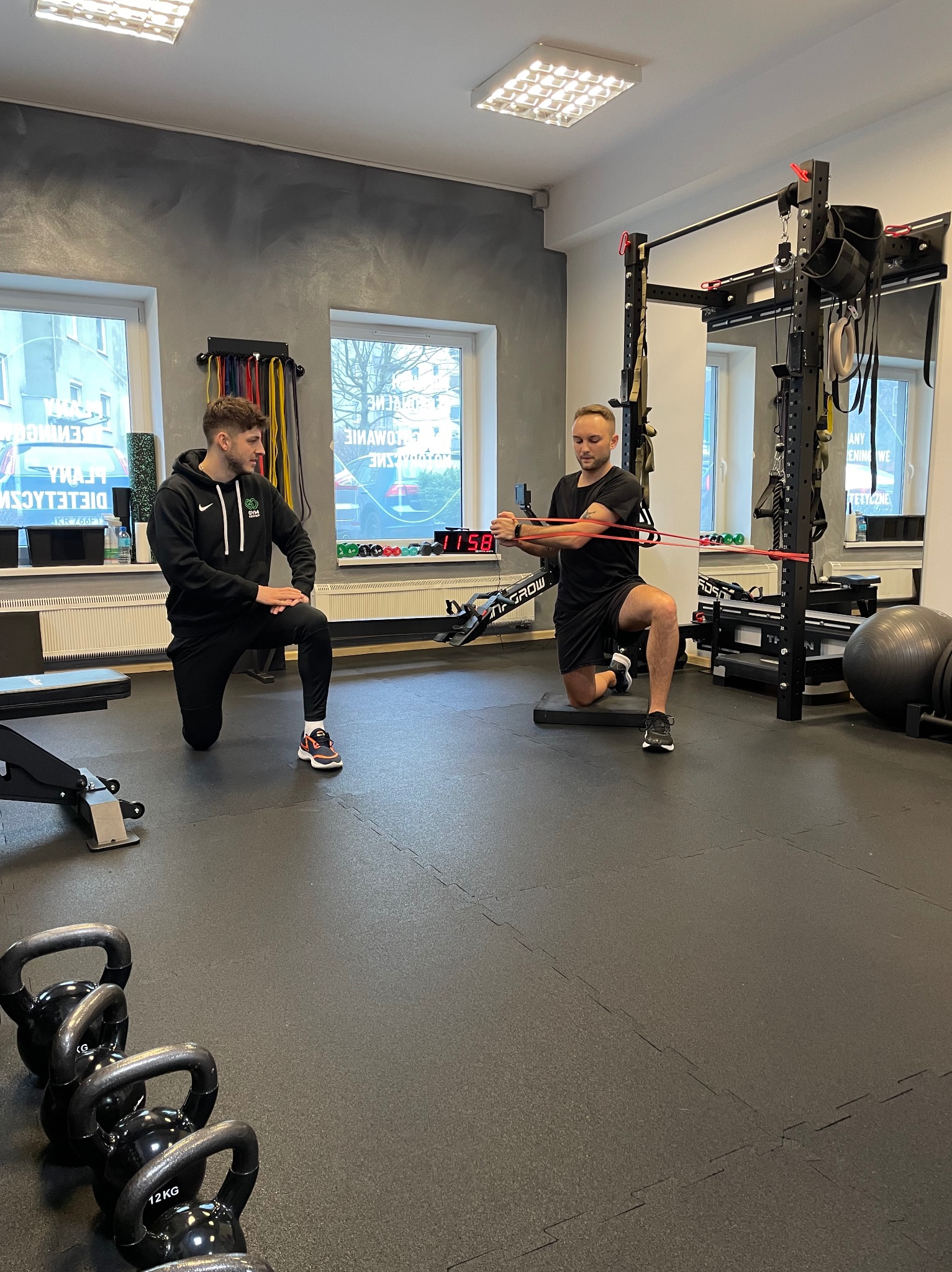 Mężczyzna ćwiczy z gumą oporową pod okiem drugiego mężczyzny w siłowni, w tle widoczne okna i sprzęt do ćwiczeń.