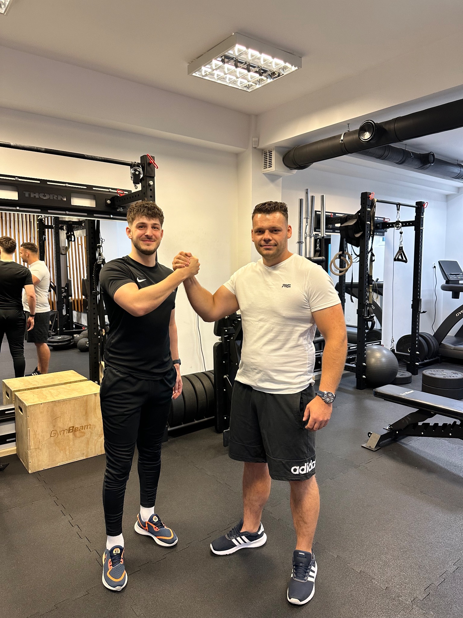 Dwóch mężczyzn w sportowych strojach ściska sobie dłonie w siłowni, w tle widoczny sprzęt do ćwiczeń siłowych i kardio.