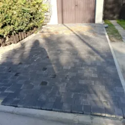 Ułożona, grafitowa kostka brukowa przed garażem z brązową bramą, widoczny cień osoby fotografującej oraz rozsypany piasek na łączeniach kostki.