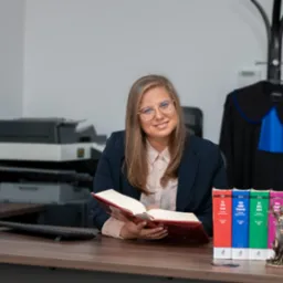 Uśmiechnięta prawniczka w okularach siedzi za biurkiem z rozłożoną księgą prawniczą, obok kodeksy i statuetka Temidy, w tle toga adwokacka