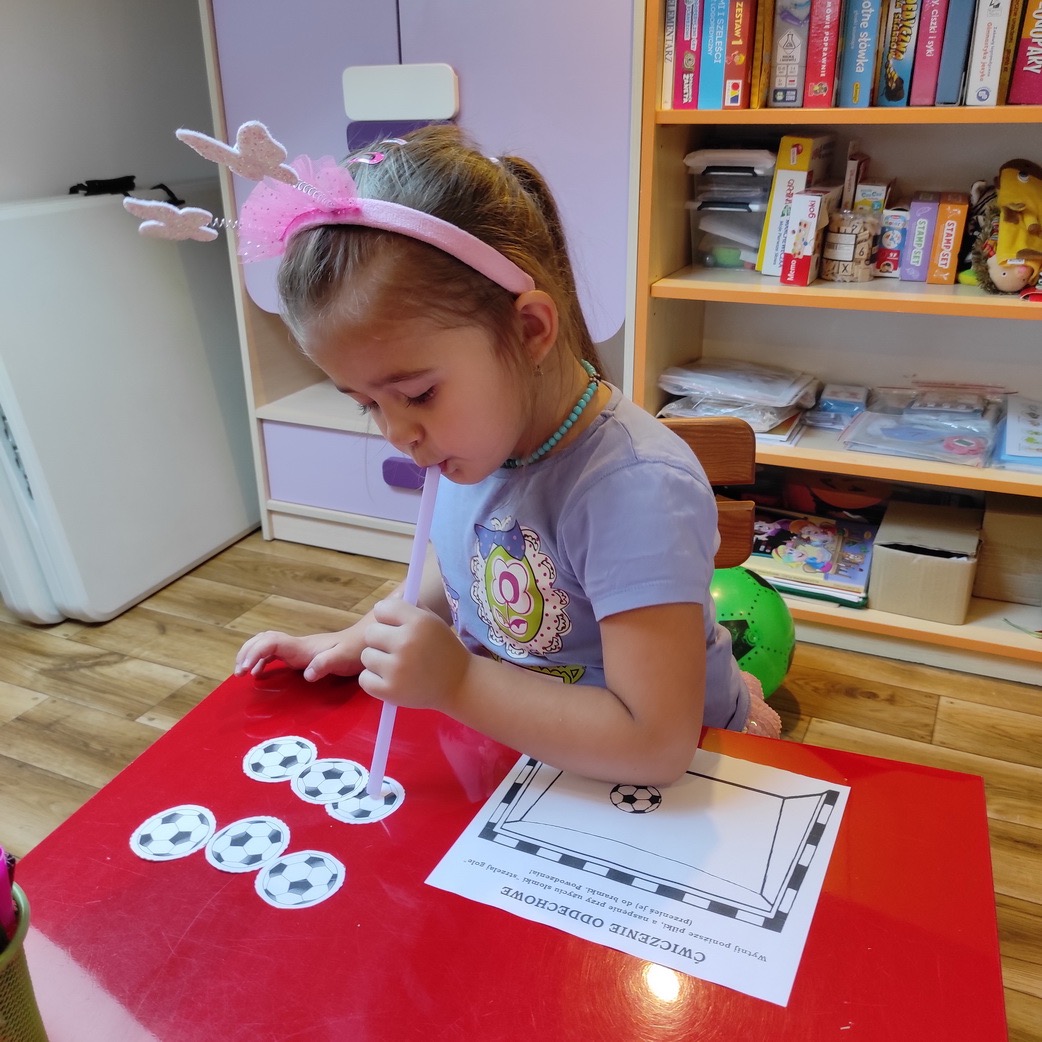 Dziewczynka z różową opaską na włosach ćwiczy oddech, dmuchając przez słomkę na papierowe piłki do bramki podczas zajęć logopedycznych.
