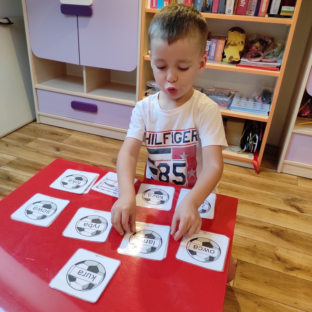 Chłopiec w białej koszulce z nadrukiem Hilfiger układa karty z obrazkami piłek i podpisanymi nazwami zwierząt i przedmiotów na czerwonym stole, ćwicząc wymowę.