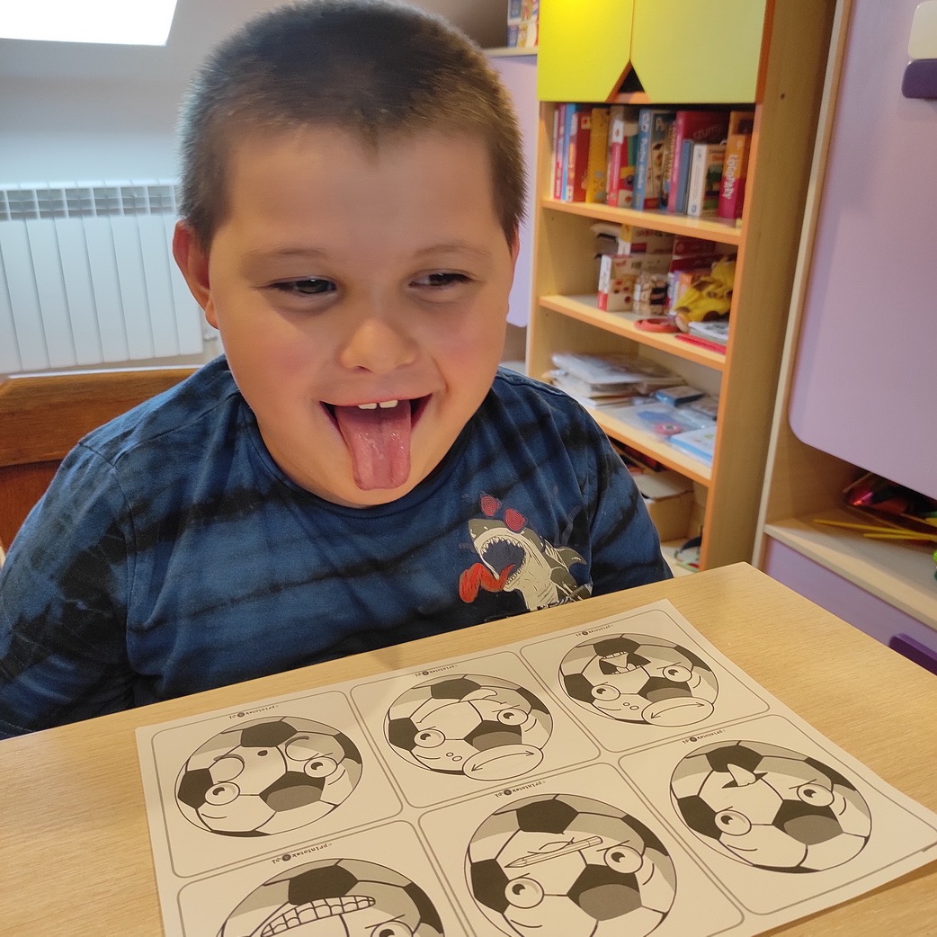 Chłopiec z wysuniętym językiem siedzi przy stole z kartką zawierającą rysunki piłek z różnymi minami, na tle regał z książkami.