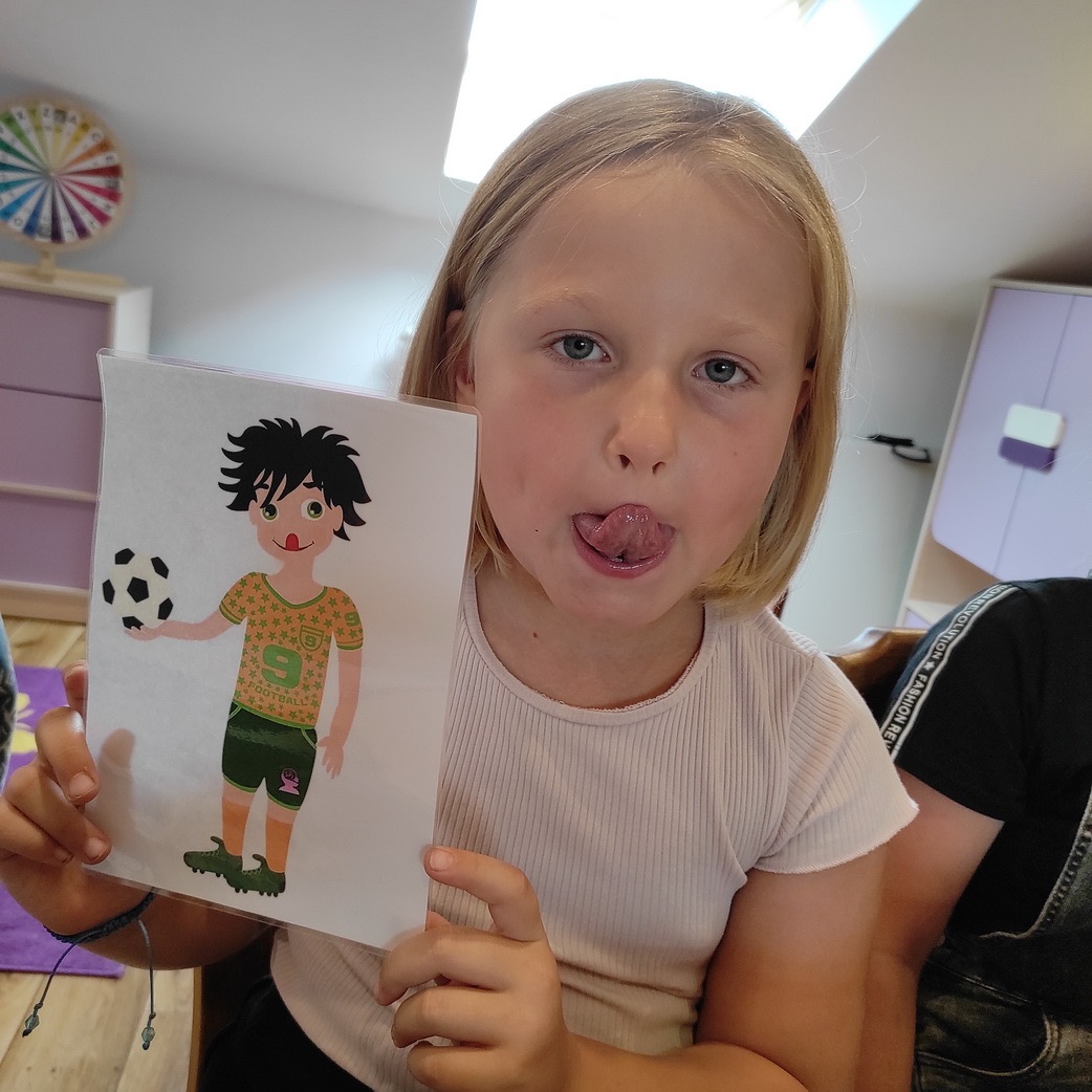 Dziewczynka z blond włosami pokazuje rysunek piłkarza z piłką, wystawiając język.