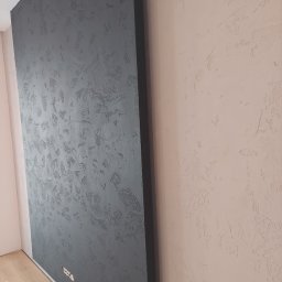 Firma usługowo-handlowa - Dwie ściany z dekoracyjnym tynkiem: szara ściana z wyraźną fakturą i odkrytymi gniazdkami elektrycznymi oraz ściana w odcieniu pudrowego różu z delikatniejszą fakturą.