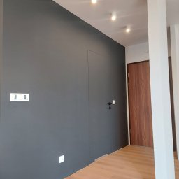 Firma usługowo-handlowa - Nowoczesne wnętrze po odświeżeniu: szara ściana z ukrytymi drzwiami, jasna podłoga z paneli, białe kolumny i oświetlenie punktowe.