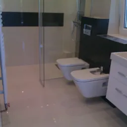 Nowoczesna łazienka z kabiną prysznicową typu walk-in, podwieszaną toaletą i bidetem oraz białą szafką z umywalką, wykończona w jasnych kolorach z ciemnymi akcentami.