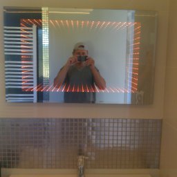 Firma usługowo-handlowa - Łazienka z podświetlanym lustrem i mozaiką, widoczny grzejnik drabinkowy i odbicie fotografa robiącego zdjęcie.