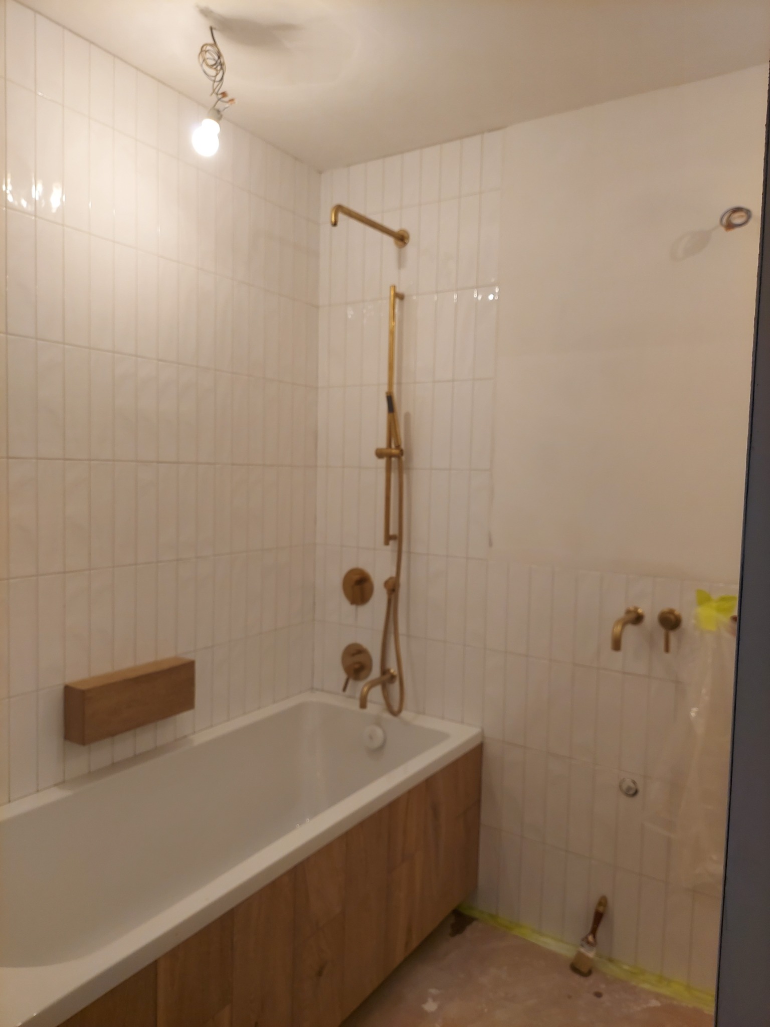 Łazienka w trakcie wykańczania: białe, wąskie płytki na ścianach, złota armatura, obudowa wanny z drewnopodobnych paneli, widoczna szczotka malarska na podłodze i zabezpieczająca folia.