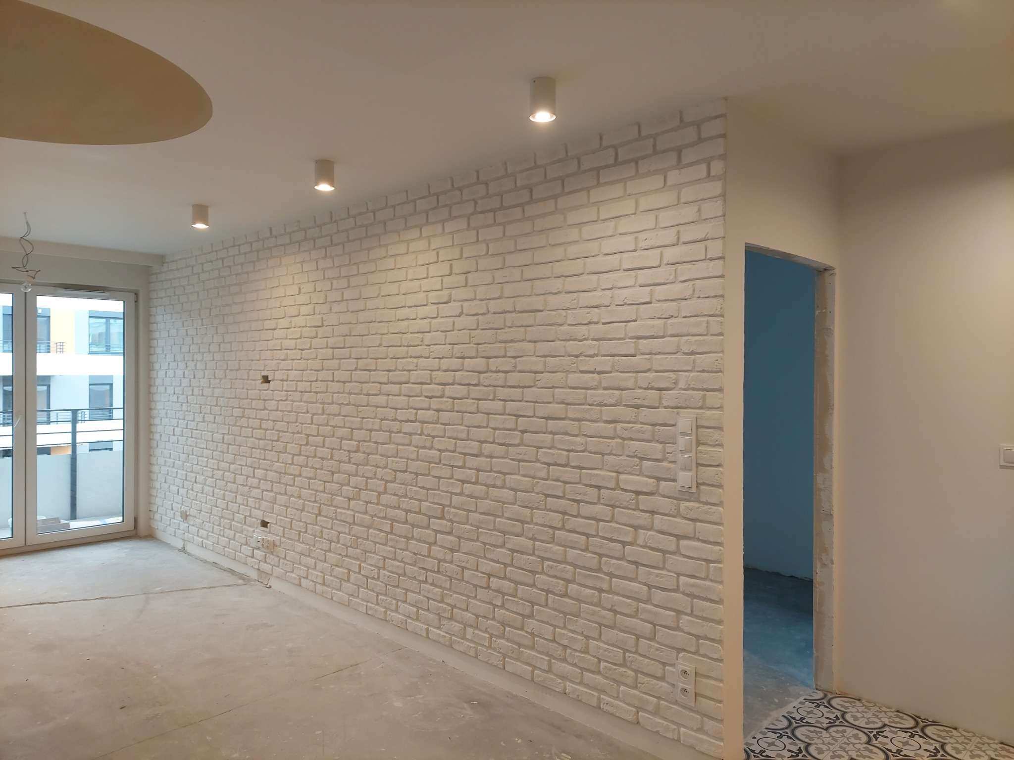 Ściana wyłożona białą cegłą dekoracyjną w nowym mieszkaniu, widoczne gniazdka elektryczne, surowa podłoga i okno balkonowe z widokiem na sąsiedni budynek.