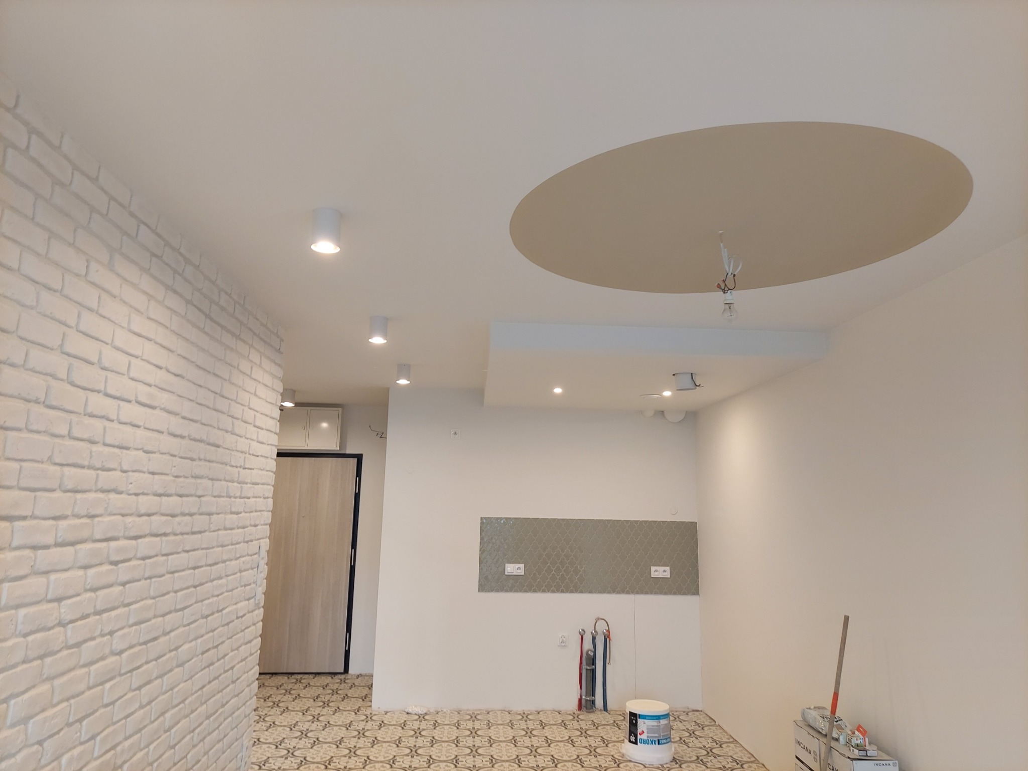 Wnętrze w trakcie odświeżania: ściana z cegły pomalowana na biało, dekoracyjny okrąg na suficie w odcieniach beżu, widoczne narzędzia malarskie i wiadro z farbą na wzorzystej podłodze.