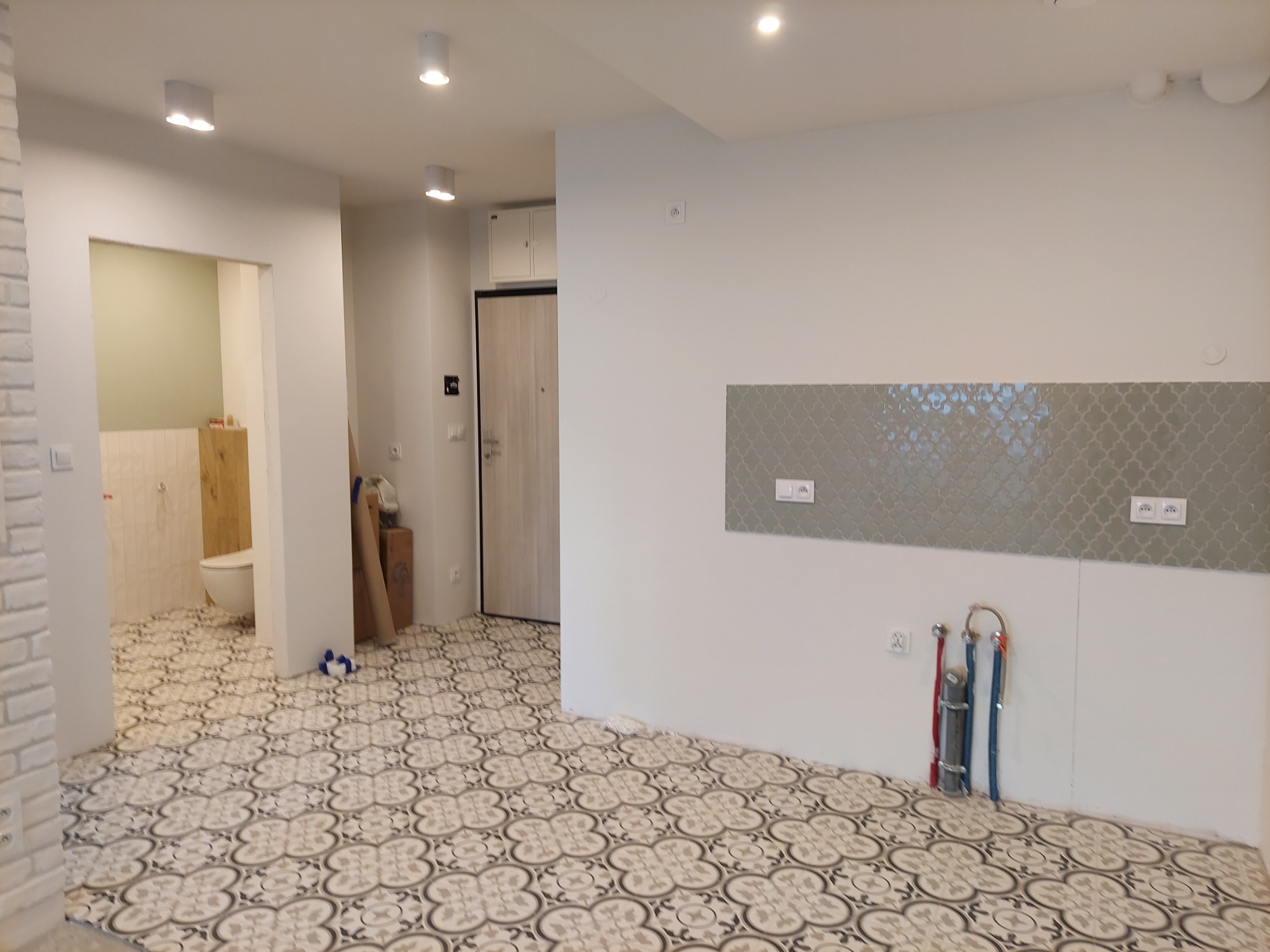 Wnętrze w trakcie remontu: wzorzysta podłoga, fragment ściany z szarą mozaiką, rury instalacyjne, widok na łazienkę z białą armaturą i okładziną ścienną.