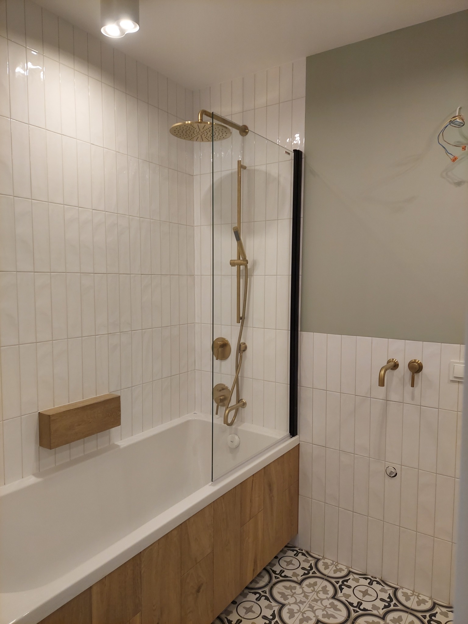 Wykończona łazienka z wanną obudowaną drewnem, przeszklonym parawanem, złotą armaturą i białymi, pionowymi płytkami na ścianach oraz wzorzystą podłogą.