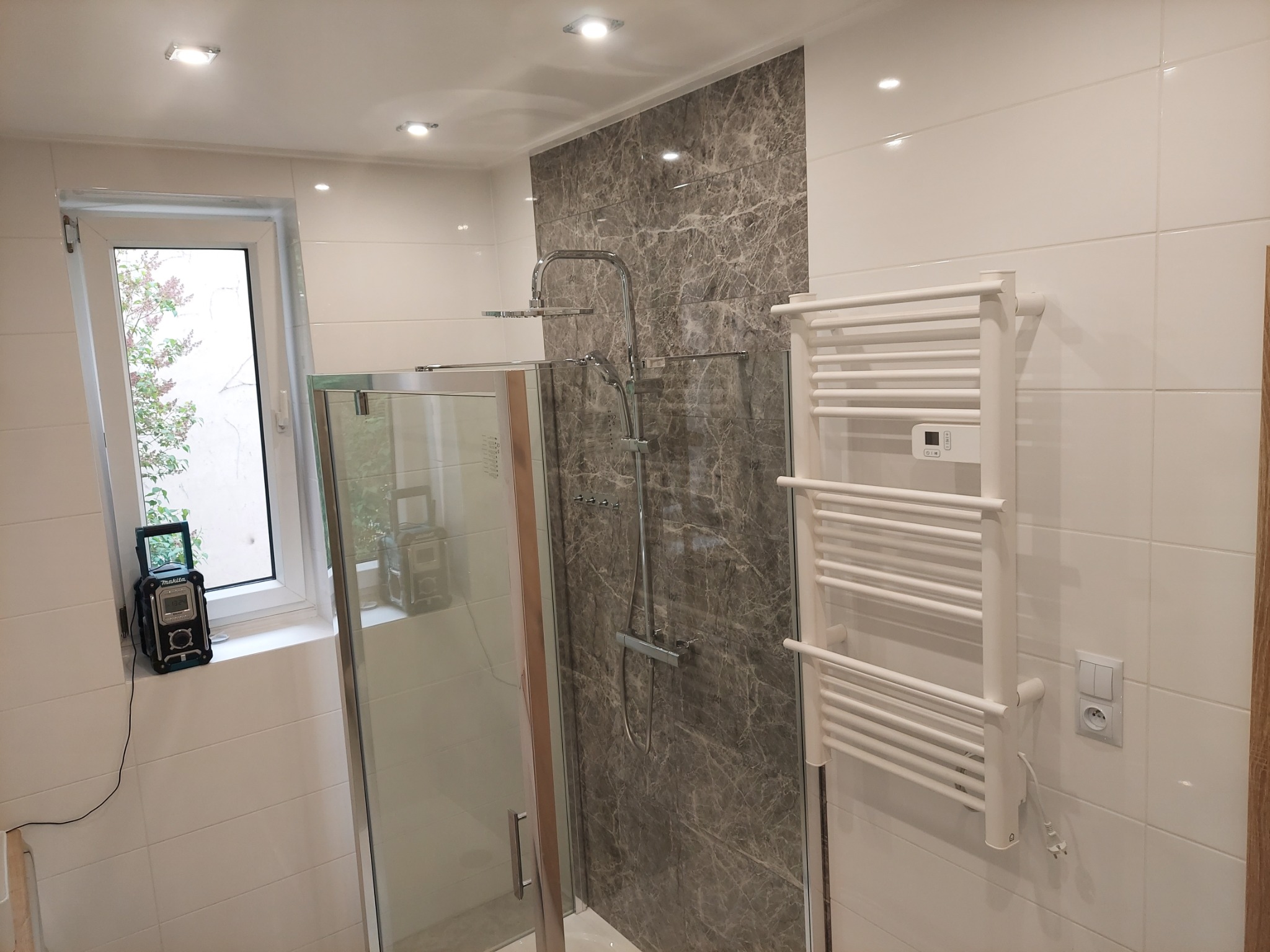 Nowoczesna łazienka z kabiną prysznicową, marmurową ścianą pod prysznicem, białymi płytkami, grzejnikiem drabinkowym i radiem budowlanym Makita na parapecie okna.