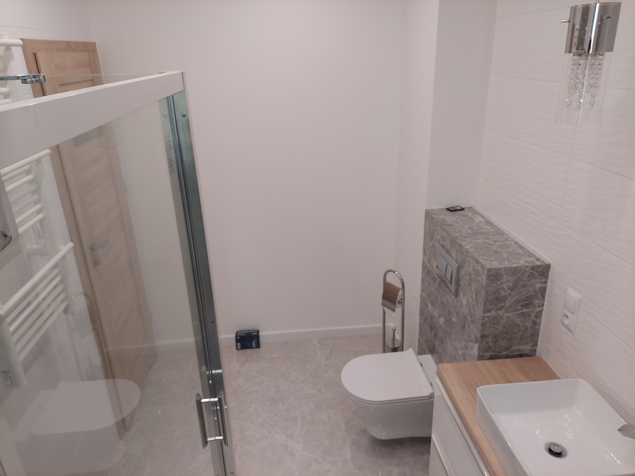 Nowoczesna łazienka z kabiną prysznicową ze szklanymi drzwiami, podwieszaną toaletą i umywalką nablatową, wykończona płytkami imitującymi marmur na podłodze i ścianie za toaletą, z ozdobnym...