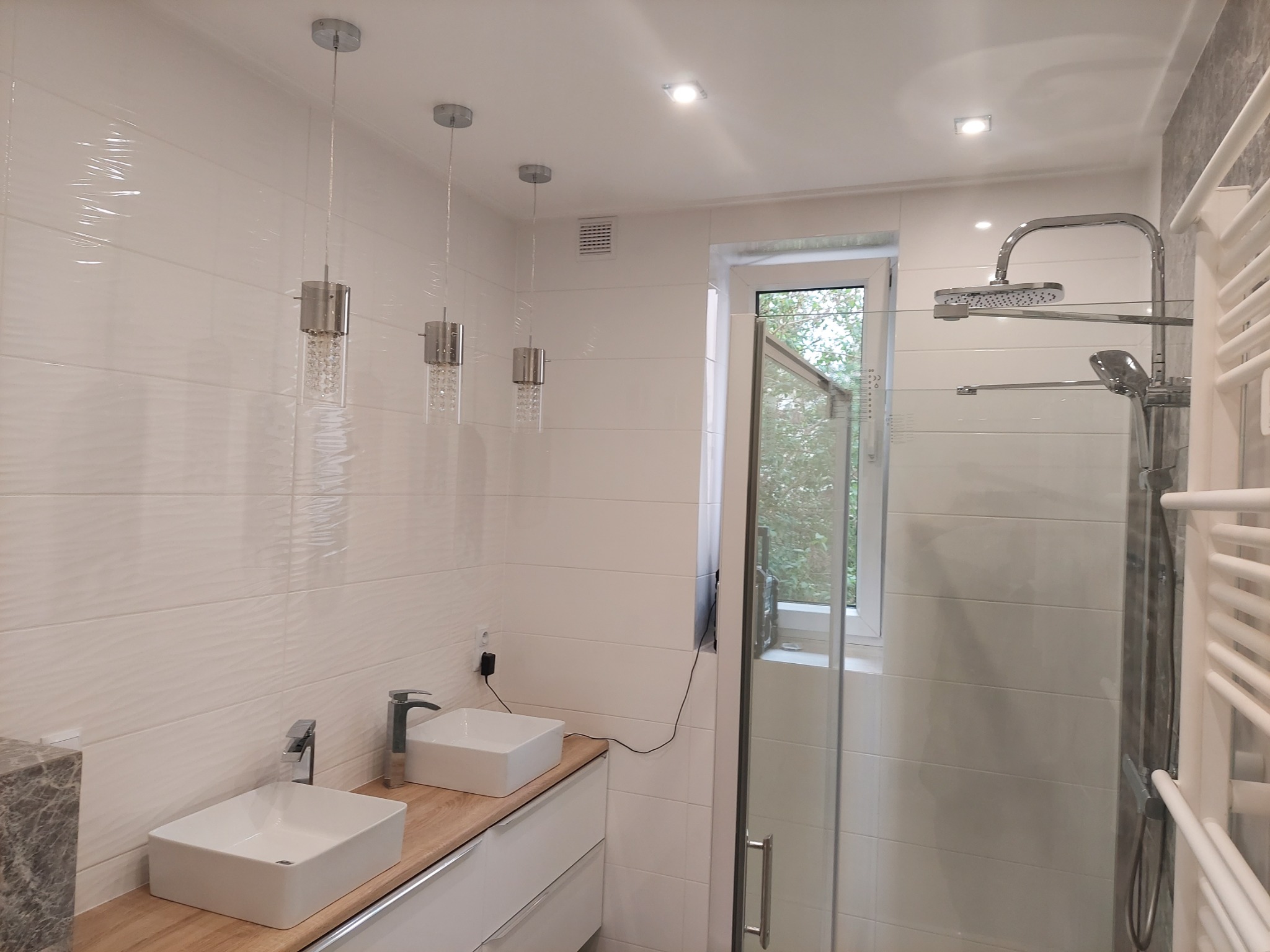 Jasna łazienka z białymi płytkami, dwiema umywalkami nablatowymi, drewnianym blatem i szklaną kabiną prysznicową z deszczownicą.