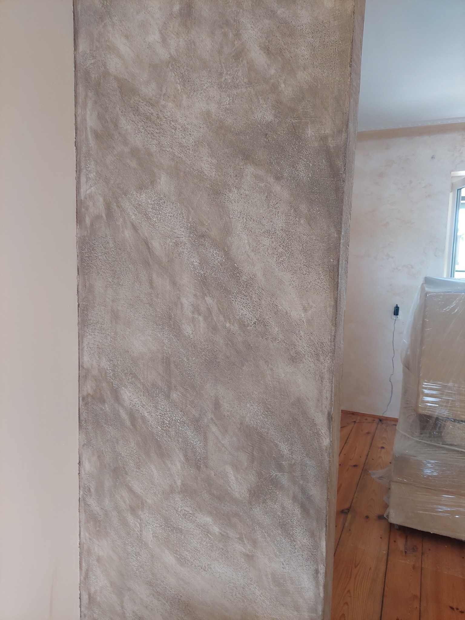 Wykończony filar z dekoracyjną, szarobeżową strukturą na ścianie, widoczne drewniane podłogi i meble zabezpieczone folią w tle.