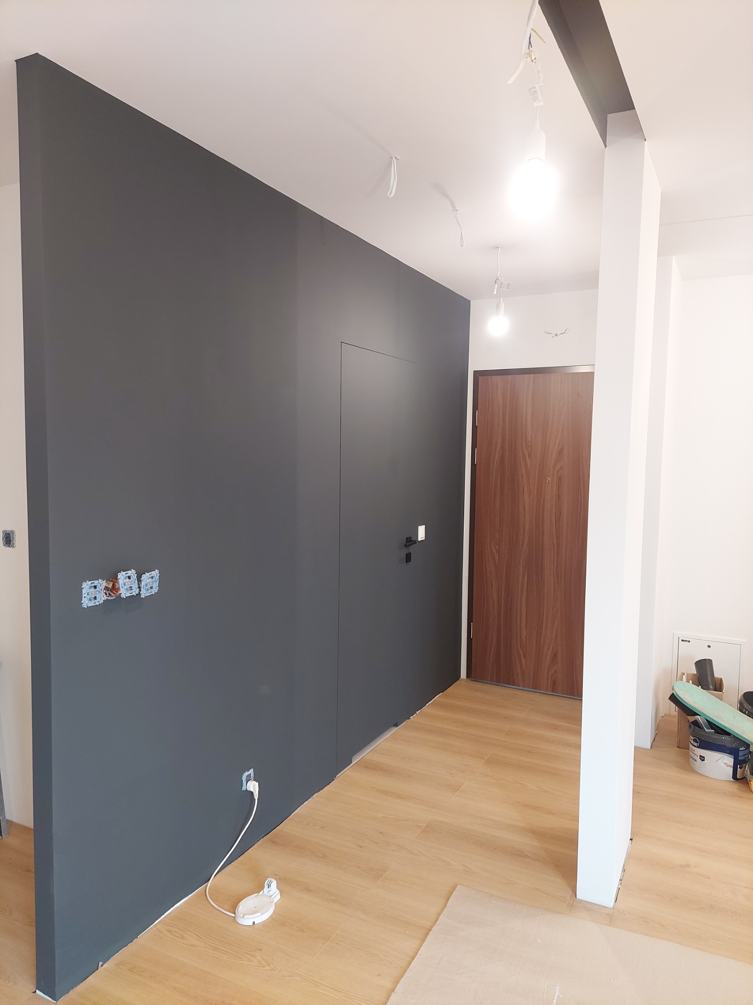 Świeżo pomalowana ściana w odcieniach szarości, z widocznymi puszkami elektrycznymi bez osprzętu i gniazdkiem z podłączonym przedłużaczem, na podłodze panele w kolorze jasnego drewna, w tle...