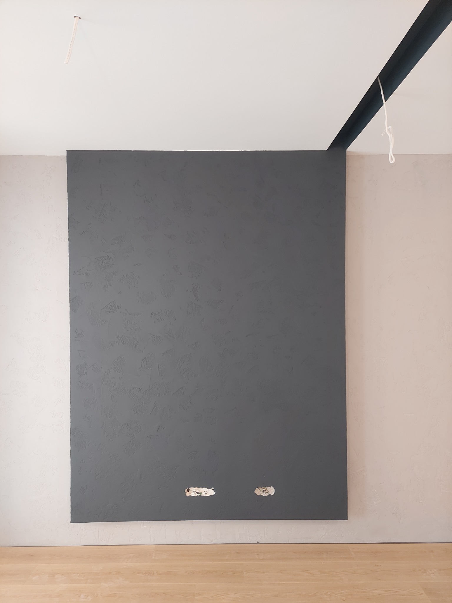 Ściana pomalowana na szaro z widoczną fakturą, obok fragment ściany w odcieniu beżu, podłoga z paneli, dwa otwory na gniazdka elektryczne w dolnej części szarej ściany.