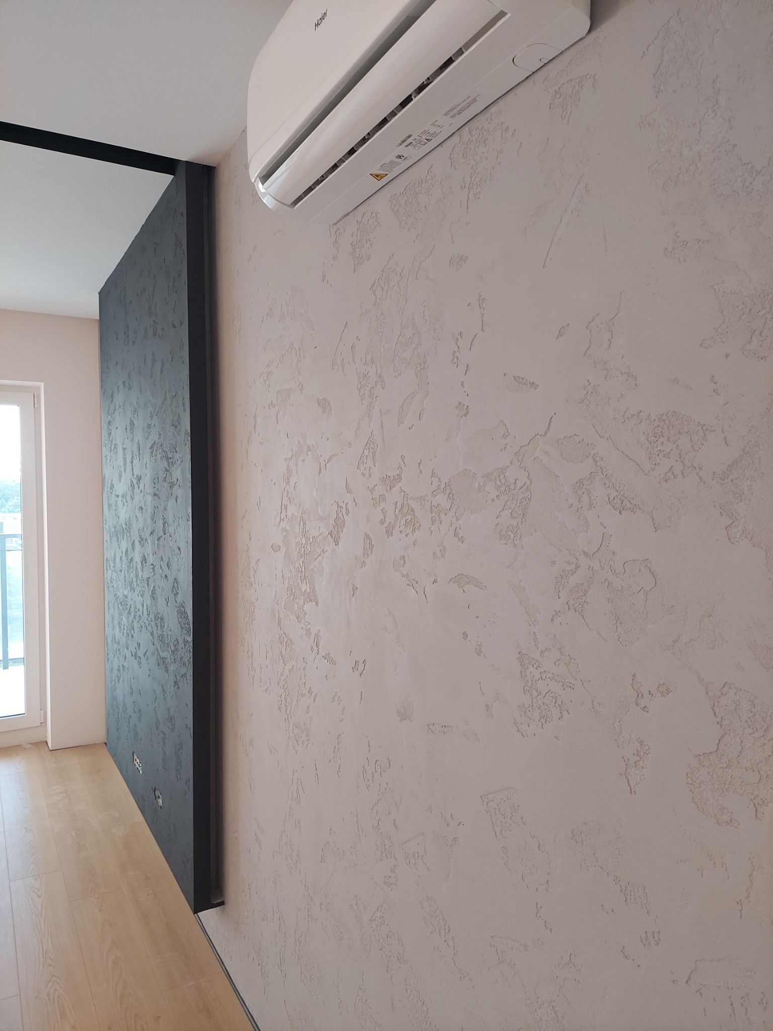 Ściana z dekoracyjnym tynkiem strukturalnym w odcieniach beżu, z zamontowanym klimatyzatorem Haier, widoczna także część ściany w kolorze ciemnoszarym i fragment podłogi z paneli.